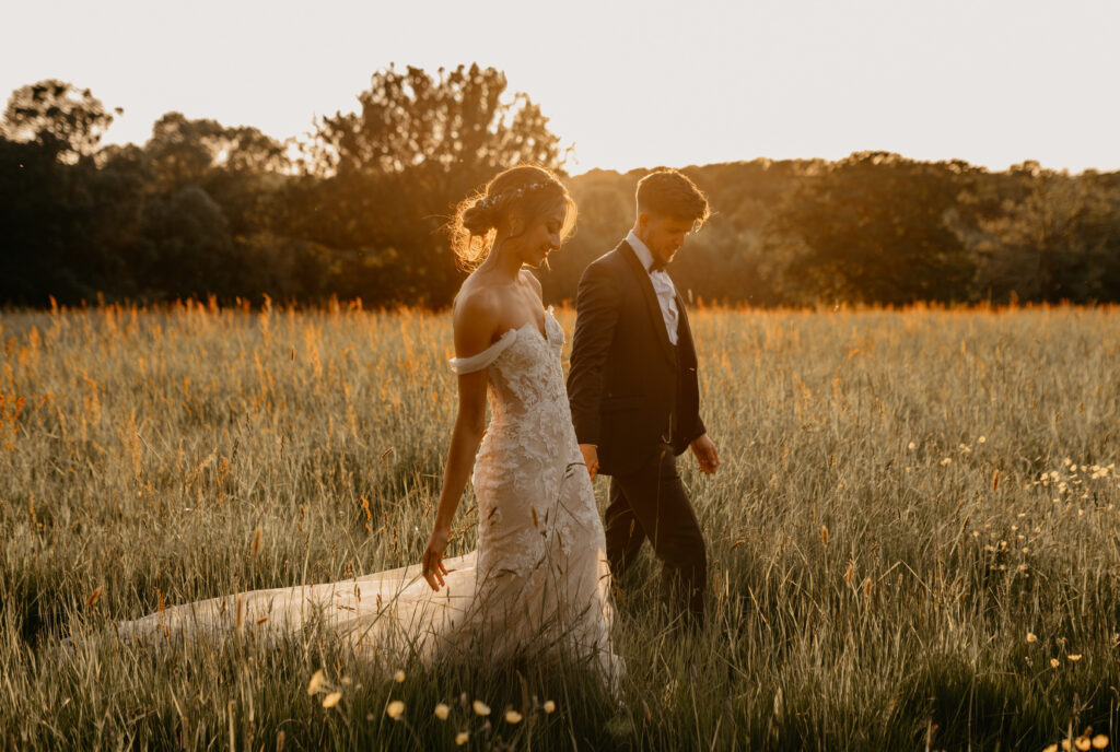 A married couple on their wedding day take a walk in a meadow at golden hour enjoying the sunset. This is at one of the uk's best wedding venues in Kent, Oak tree barn weddings.